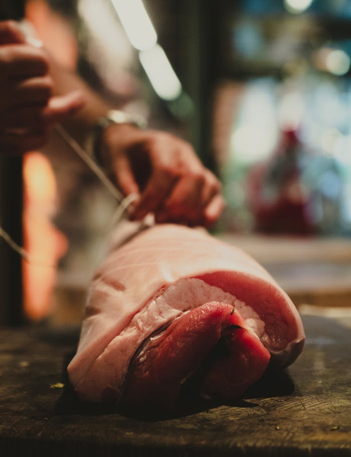who-we-are-02 A close-up view of a raw pork cut being tied with string by hands in a warm, inviting kitchen setting.
