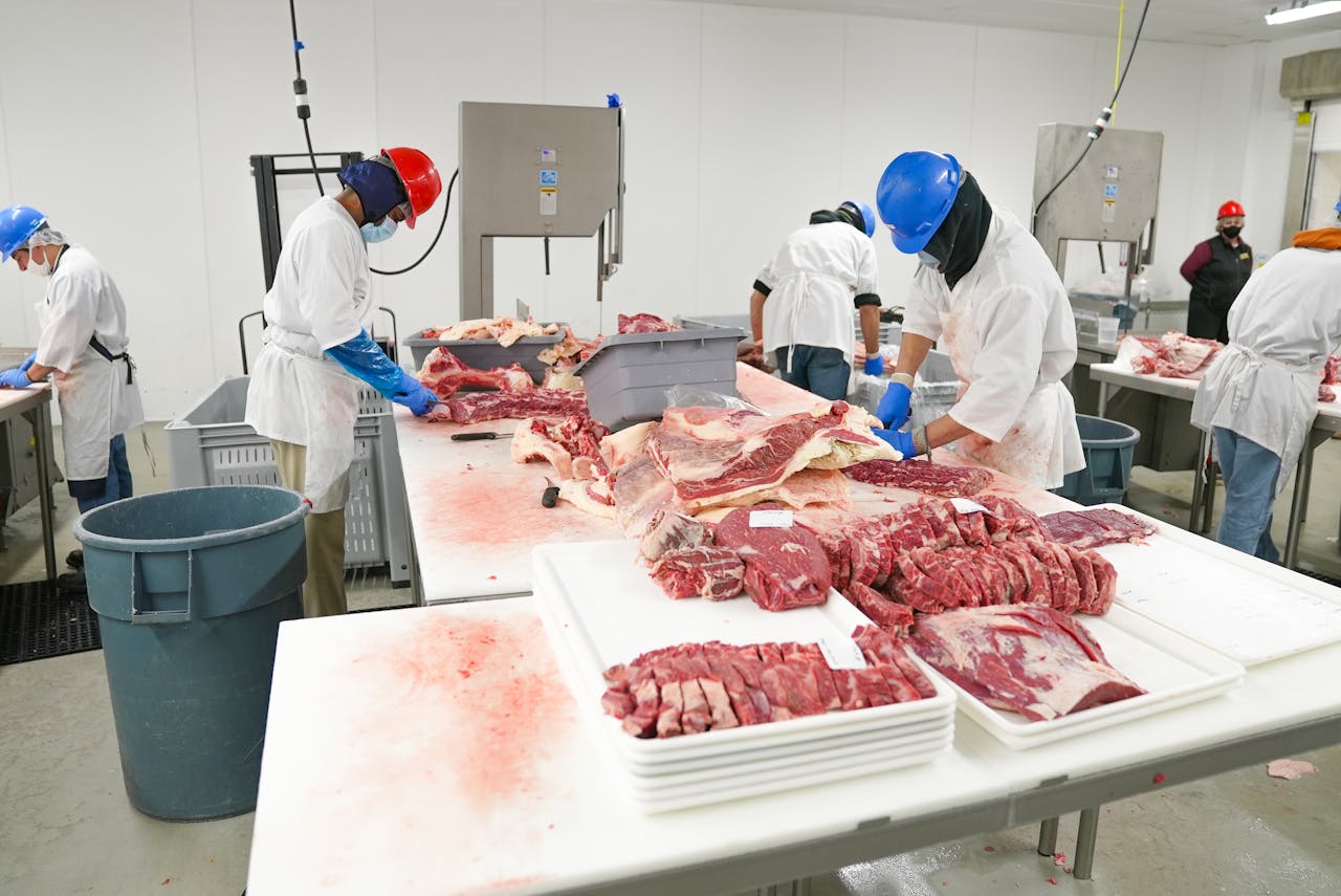 about-03 Workers in a meat processing facility wearing safety gear and cutting meat indoors.